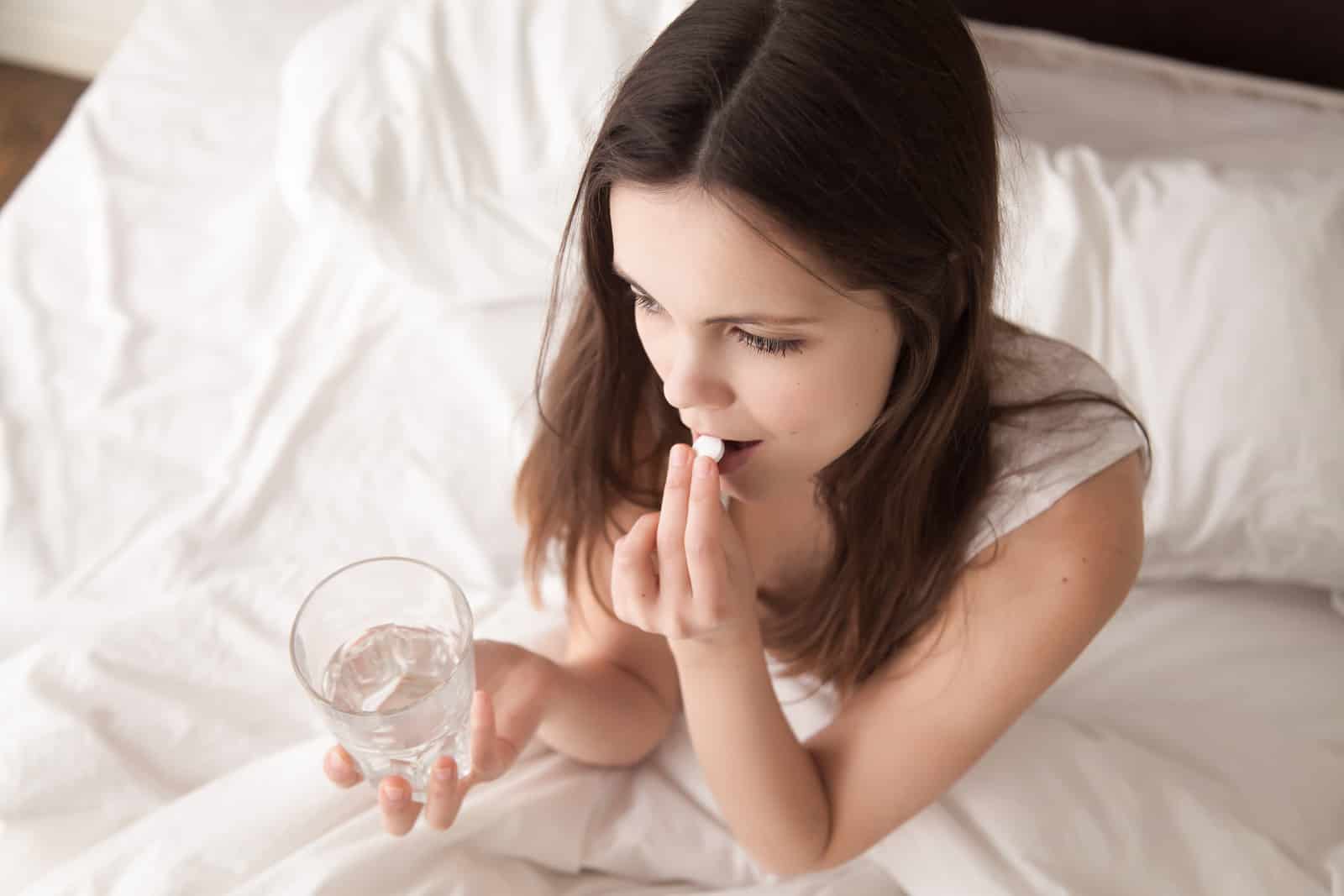 women and drug addiction - Stressed woman drinking white round pill while sitting in bed with glass of water in hand.