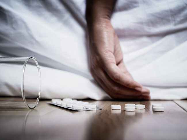 Hand hanging off bed with empty glass and pills on floor, illustrating oxymorphone overdose