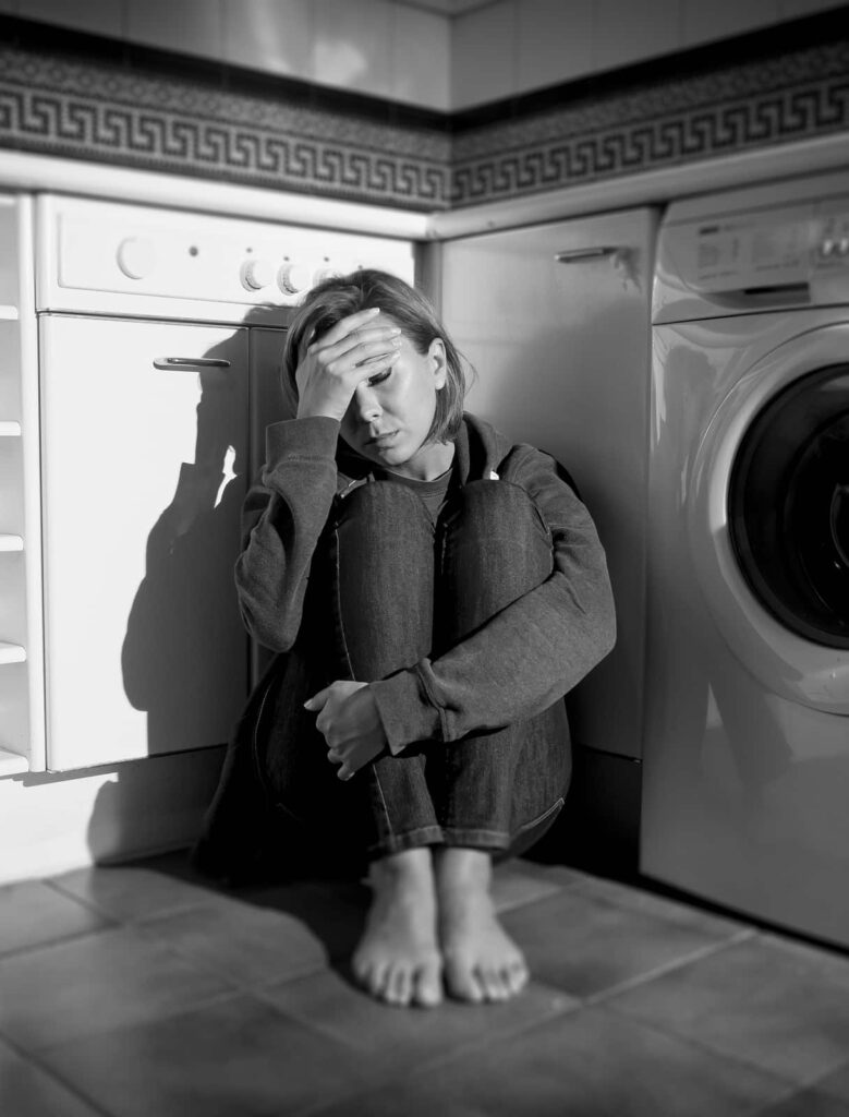 lonely depressed and sick middle aged woman sitting alone on kitchen floor in stress depression and sadness feeling miserable in barefoot looking desperate in black and white vertical composition
