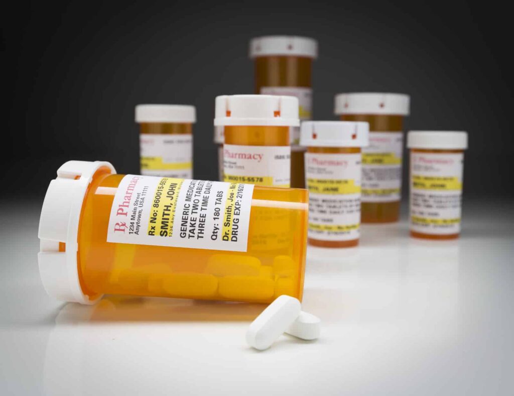 Variety of Non-Proprietary Medicine Bottles and Large Pills on Reflective Surface With Grey Background.