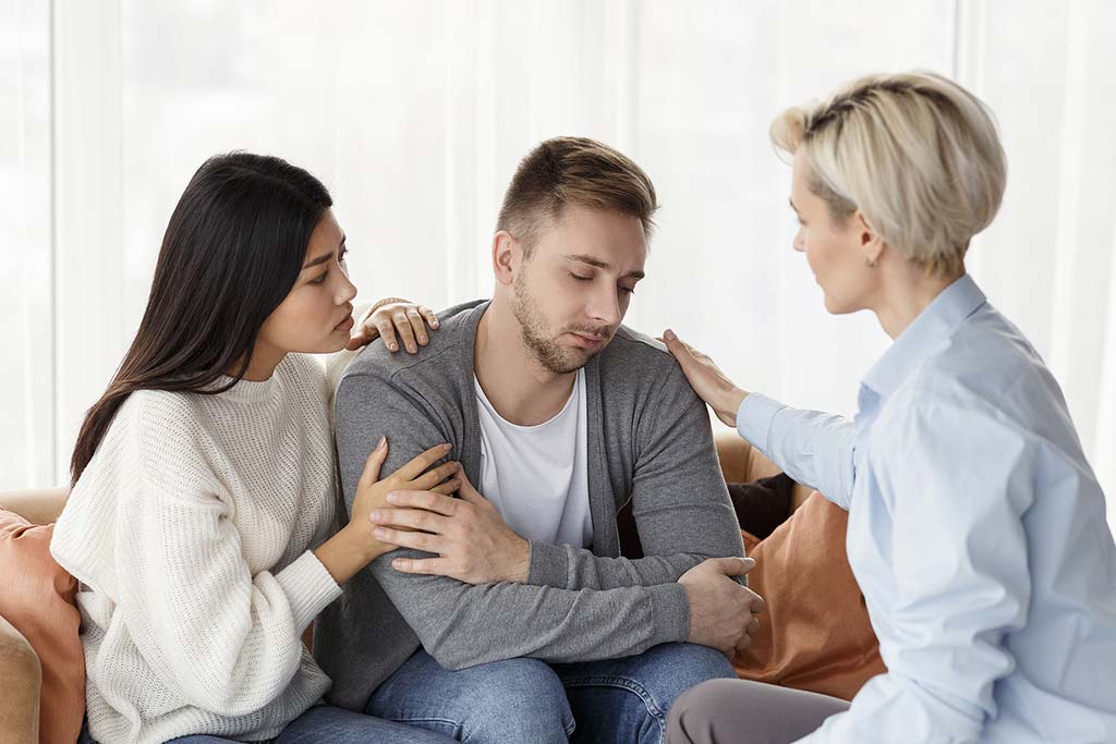 Concept of asking for help in addiction recovery. Photo of wife and family psychologist comforting and supporting depressed addicted husband at psychotherapy session indoor.