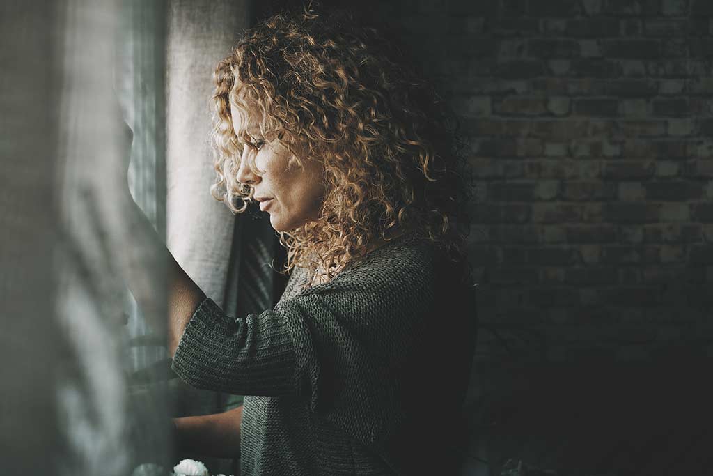 Photo of a sad, lonely woman inside her home, looking out the window - loneliness, isolation concept