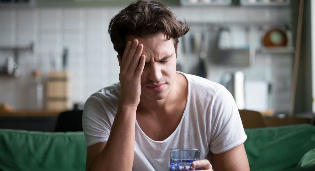 Young man suffering from strong headache or migraine sitting with glass of water in the kitchen, millennial guy feeling intoxication and pain touching aching head, alcohol withdrawal concept